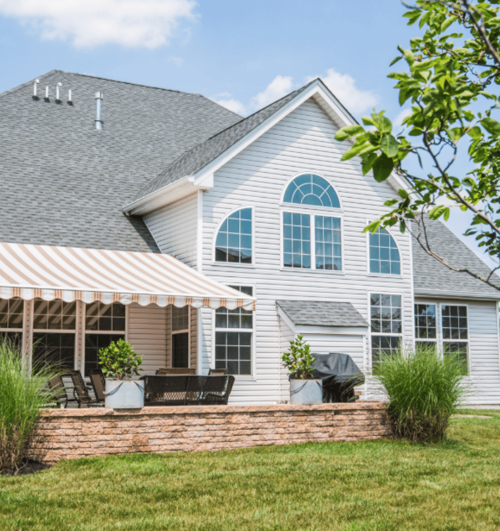 White two-story house with stone patio featuring a striped retractable awning, outdoor seating, and landscaping; Sunesta 45+ years made in America badge in upper left corner