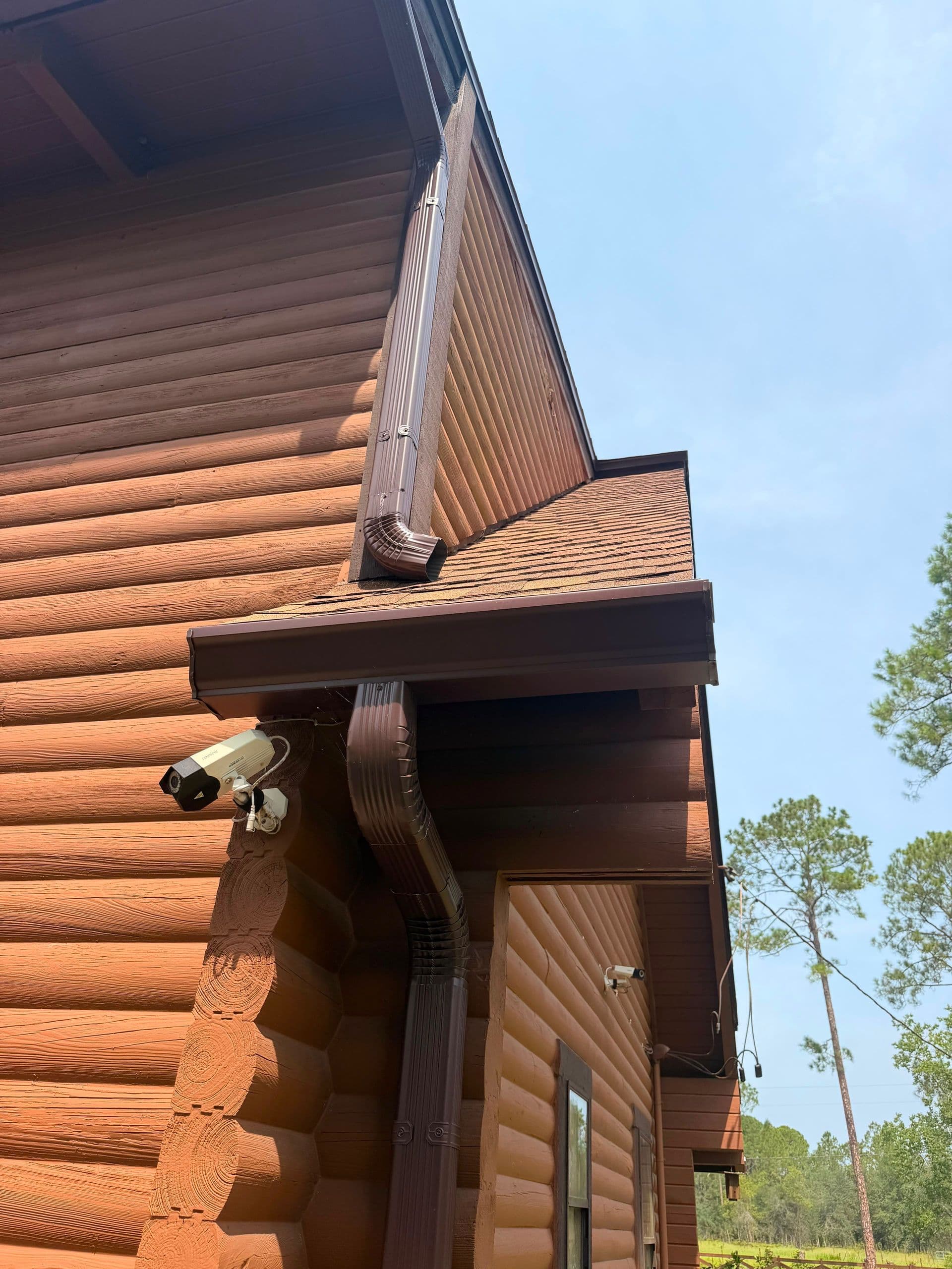Exterior corner of log cabin with brown metal gutters and downspout, security camera mounted above doorway, surrounded by tall pine trees