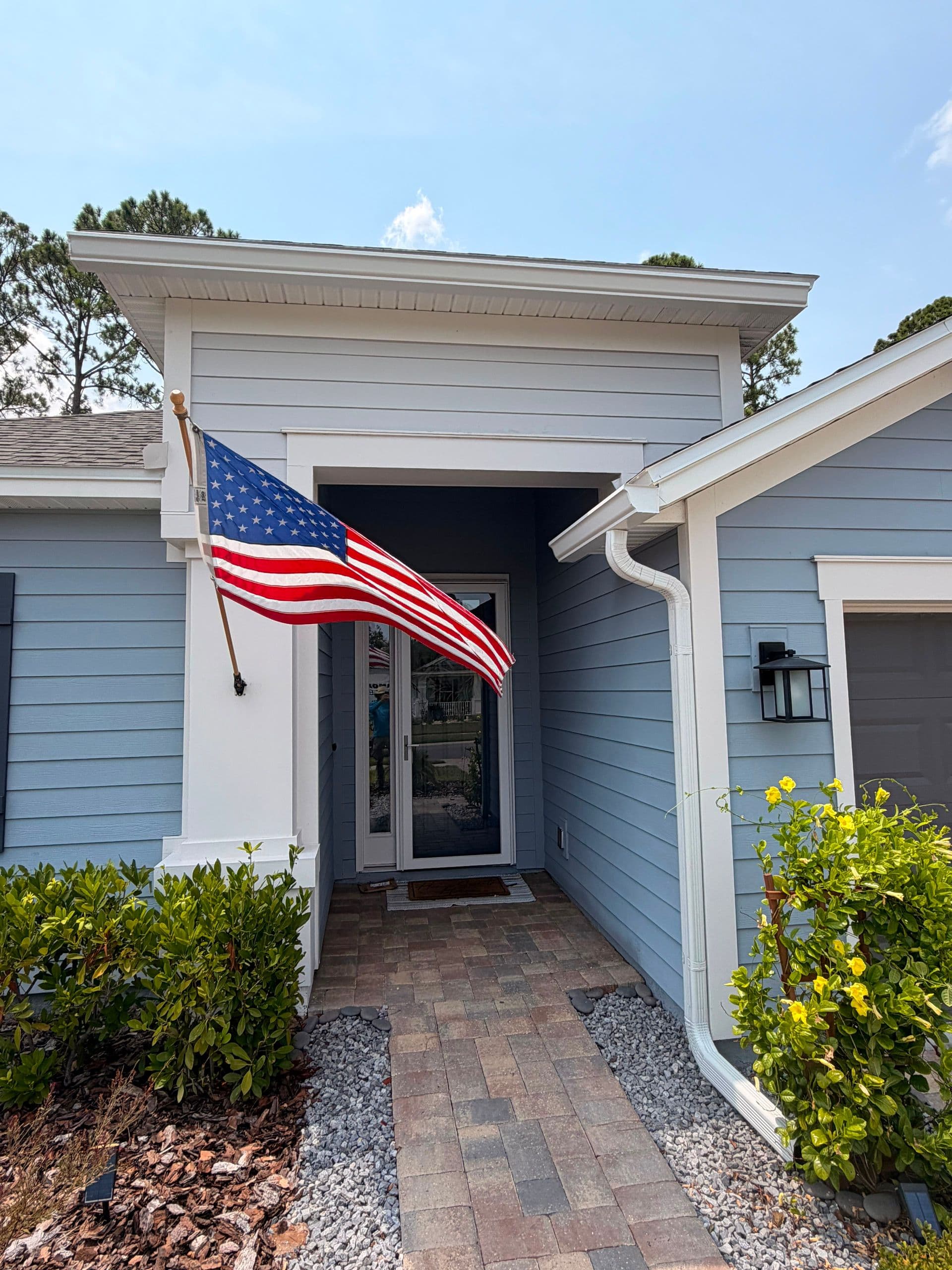 Modern light blue and white house with American flag hanging from entrance, brick pathway, yellow flowering shrubs, and attached garage