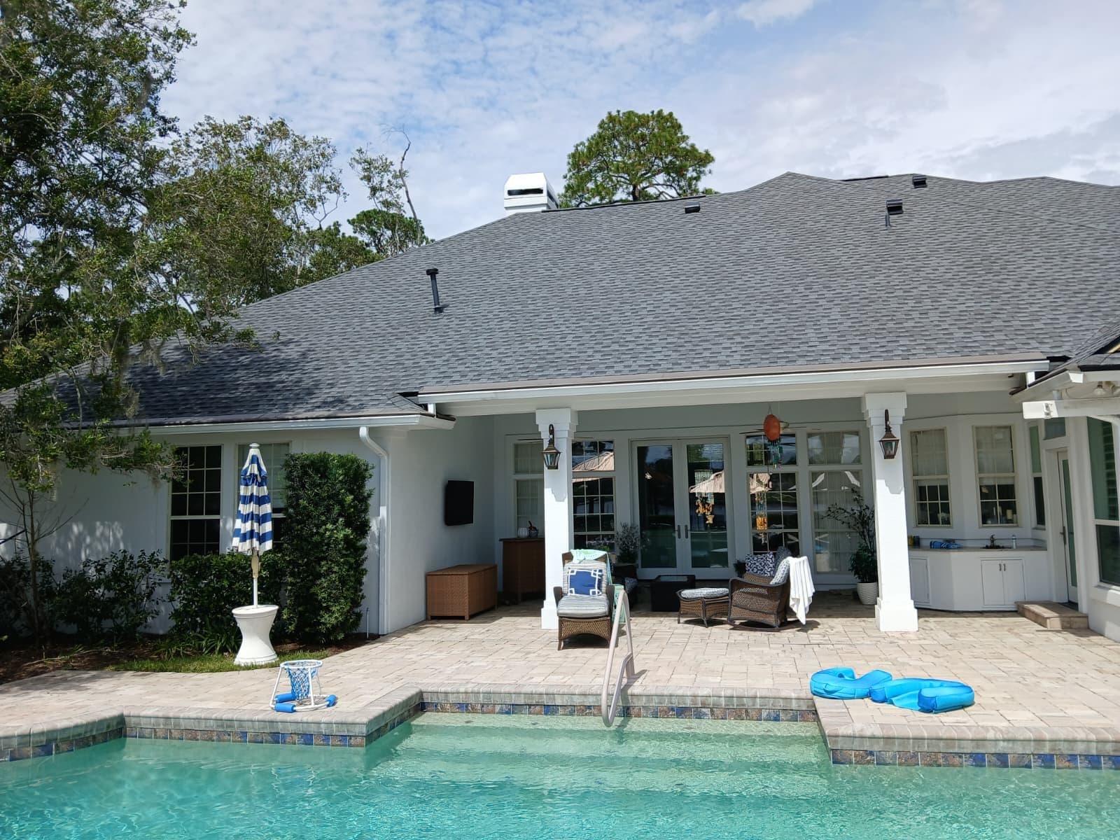 White residential home with gray roof and covered patio overlooking pool, featuring loungers and blue striped umbrella on paved deck