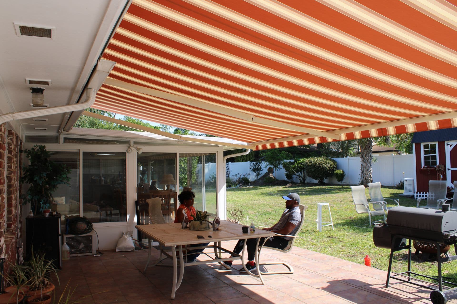 Retractable awning with orange and white stripes extends over a backyard patio, providing shade for people relaxing at a dining table near glass doors to the house