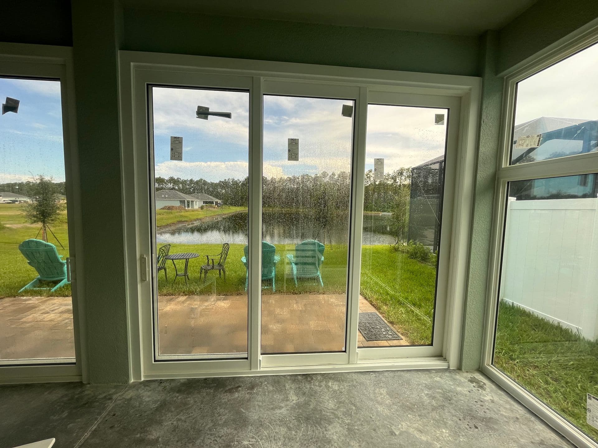 View through large sliding glass doors from interior room overlooking grassy backyard with pond, green Adirondack chairs, and treeline under cloudy sky