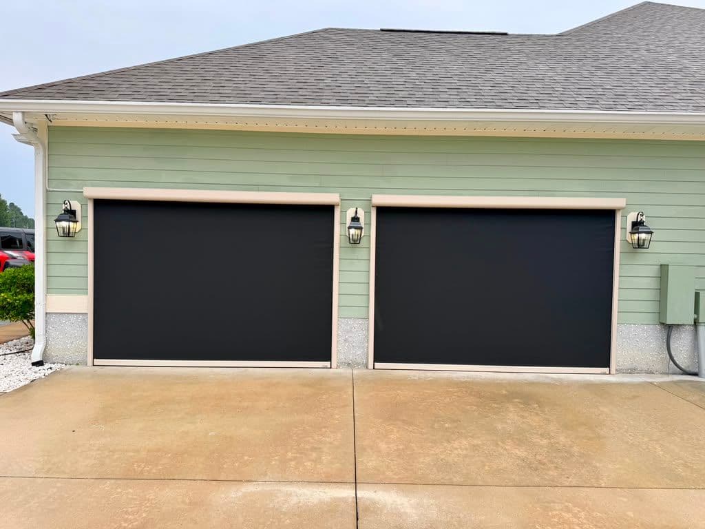 Two-car garage with black panel doors and tan trim attached to green and white siding house with lantern lighting fixtures on either side
