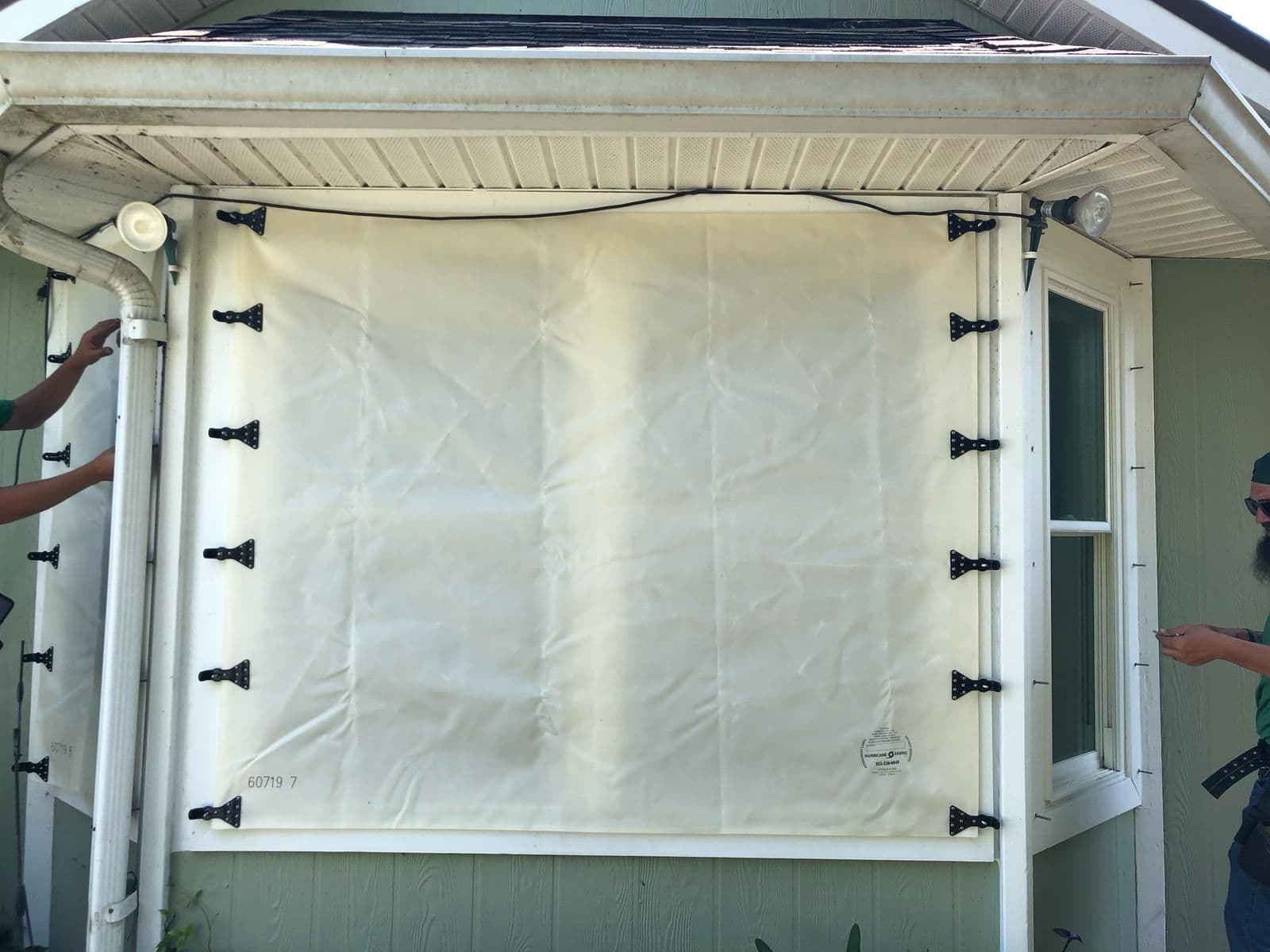 Retractable shade screen installed on porch or patio with black hardware clips securing cream-colored fabric to white frame