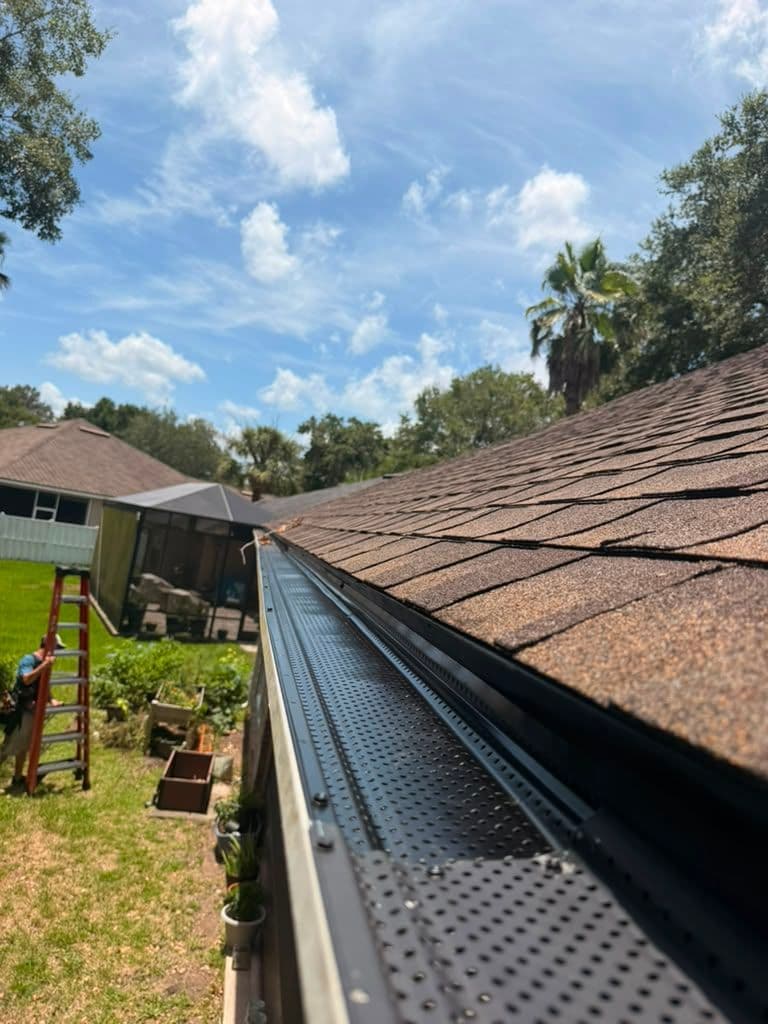 Close-up of a metal gutter guard installed along the roof edge of a residential home with a yard and shed visible below