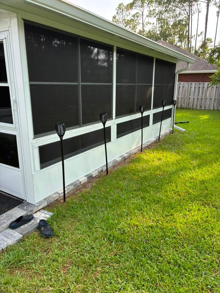 Screened porch addition with black mesh panels and white frame attached to house, featuring concrete foundation and metal support posts in grassy yard