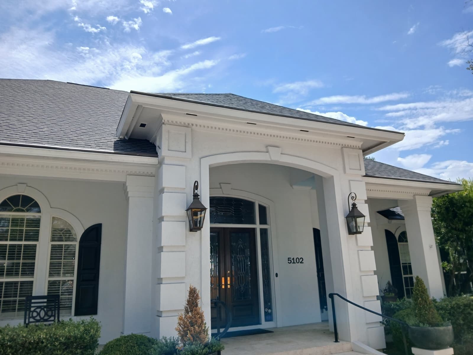 Modern white residential home with dark roof, arched entry portico, blue front door marked 5102, black lantern sconces, and manicured landscaping under blue sky