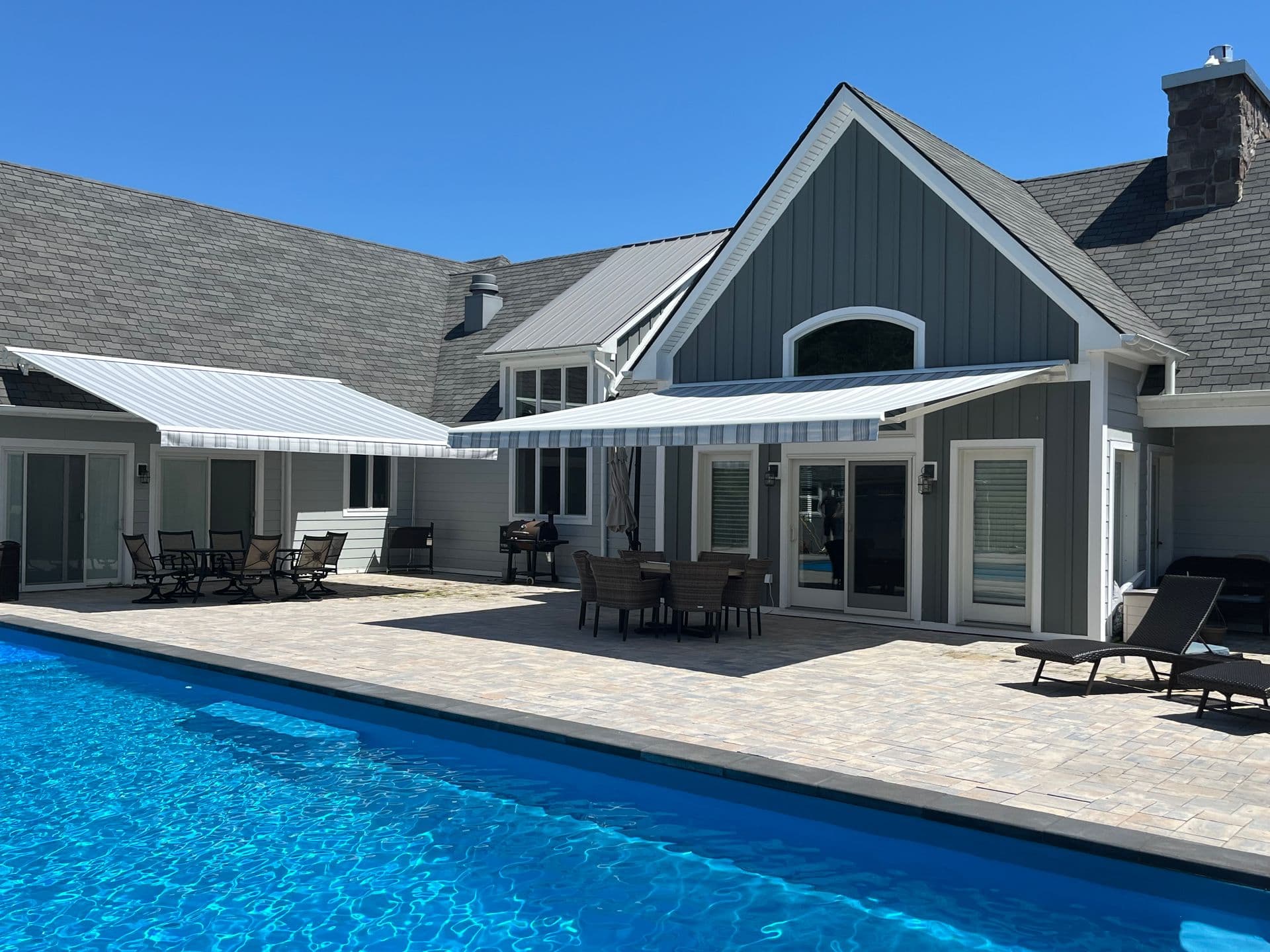 Modern residential home with dark gray and white exterior, metal roof, and covered patio overlooking an in-ground swimming pool with paved deck and outdoor lounge seating