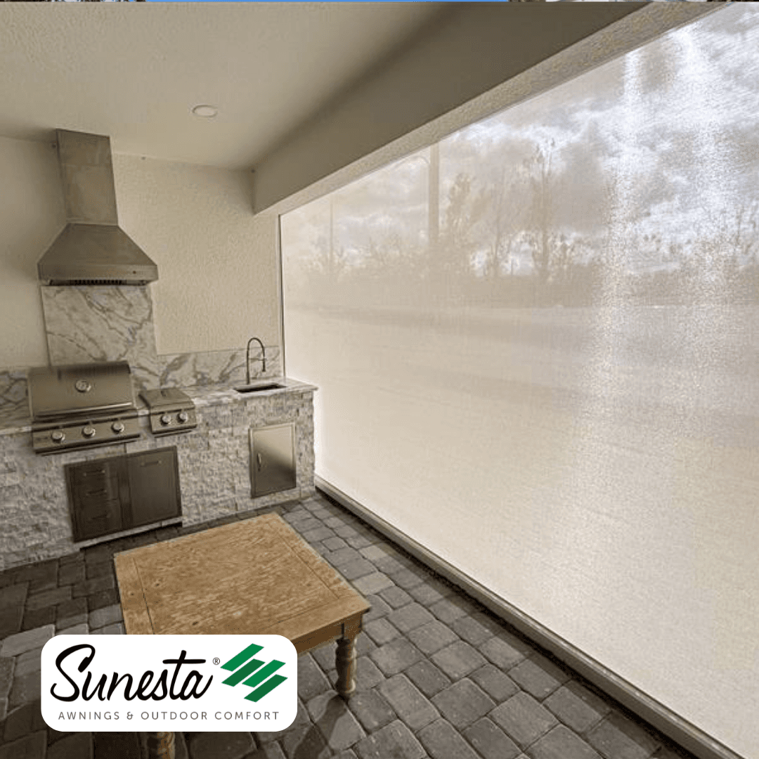 Outdoor kitchen patio with Suresta retractable shade screen deployed across large window, showing stainless steel grill, hood vent, and marble backsplash
