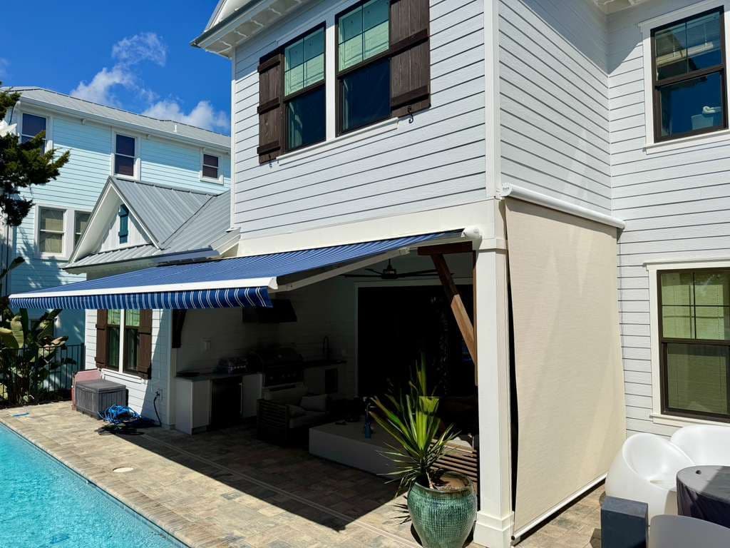 Residential home with blue and white striped retractable awning over covered patio area, adjacent to swimming pool, with white siding and dark-trimmed windows