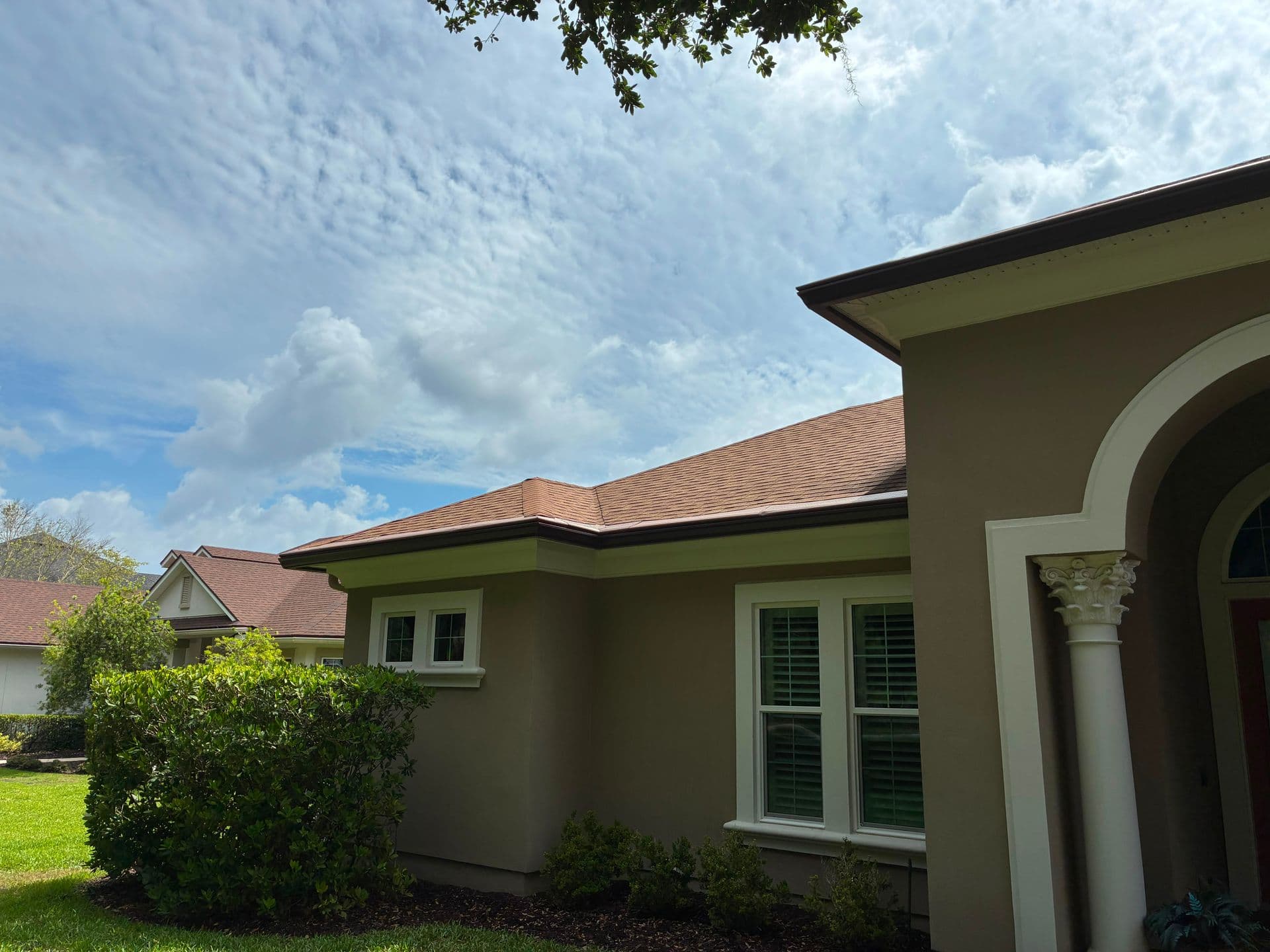 Tan stucco single-story home with tile roof, white-trimmed windows and arched entryway, surrounded by landscaped shrubs and green lawn